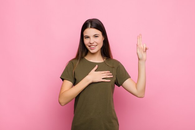 Foto mujer bonita joven que parece feliz, confiada y confiable, sonriendo y mostrando el signo de la victoria, con una actitud positiva contra la pared rosa