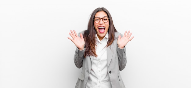 Mujer bonita joven que parece feliz y emocionada, sorprendida con una sorpresa inesperada con ambas manos abiertas al lado de la pared blanca aislada