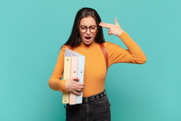 Mujer bonita joven que parece infeliz y estresada, gesto de suicidio haciendo un signo de pistola con la mano, apuntando a la cabeza. concepto de estudiante