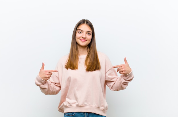 Mujer bonita joven que parece orgullosa, arrogante, feliz, sorprendida y satisfecha, apuntando a sí misma, sintiéndose como un ganador sobre la pared blanca