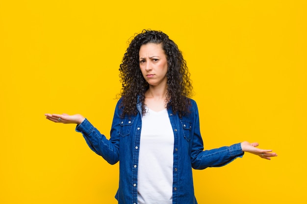 Foto mujer bonita joven que parece perpleja, confundida y estresada, preguntándose entre diferentes opciones, sintiéndose insegura contra la pared naranja