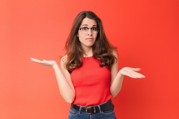 Foto mujer bonita joven que parece perpleja, confundida y estresada, preguntándose entre diferentes opciones, sintiéndose insegura sobre la pared roja