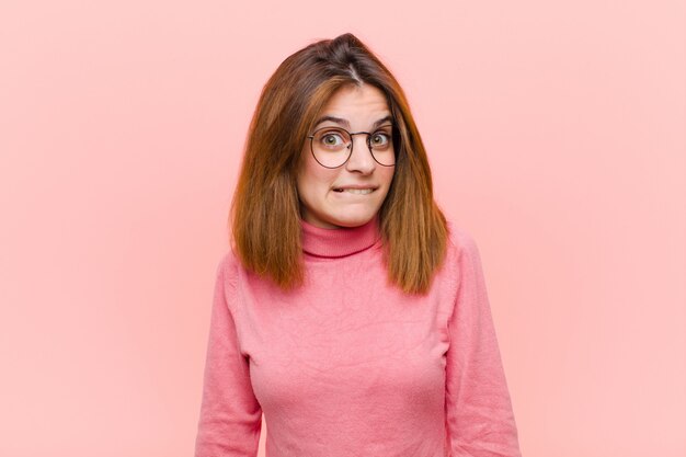 Mujer bonita joven que parece perpleja y confundida, mordiendo el labio con un gesto nervioso, sin saber la respuesta al problema contra la pared rosa