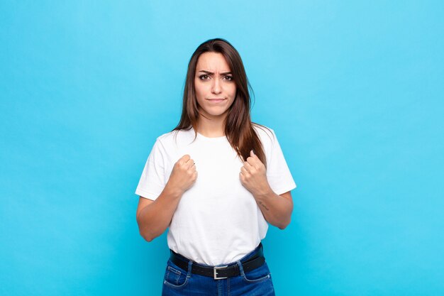 Mujer bonita joven que parece segura, enojada, fuerte y agresiva, con los puños listos para pelear en posición de boxeo contra la pared azul