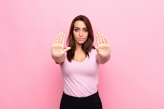 Mujer bonita joven que parece seria, infeliz, enojada y disgustada, prohibiendo la entrada o diciendo que pare con ambas palmas abiertas sobre la pared rosa