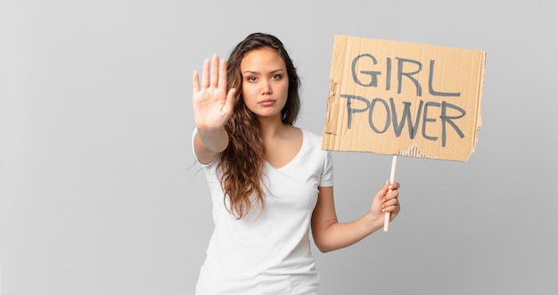 Mujer bonita joven que parece seria mostrando la palma abierta haciendo gesto de parada y sosteniendo una pancarta de poder femenino