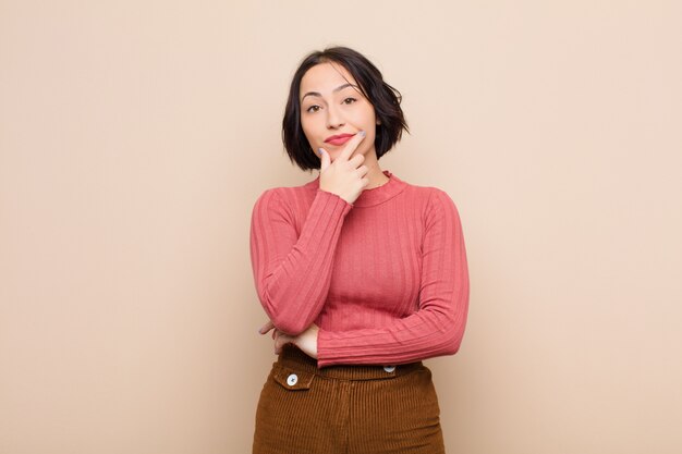 Mujer bonita joven que parece seria, reflexiva y desconfiada, con un brazo cruzado y la mano en la barbilla, opciones de ponderación sobre la pared beige