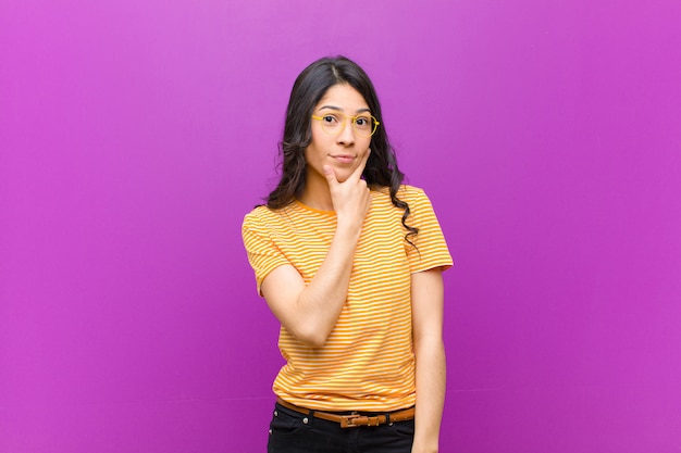 Mujer bonita joven que parece seria, reflexiva y desconfiada, con un brazo cruzado y la mano en la barbilla, opciones de ponderación sobre la pared púrpura