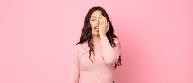 Mujer bonita joven que parece somnolienta, aburrida y bostezando, con dolor de cabeza y una mano cubriendo la mitad de la cara