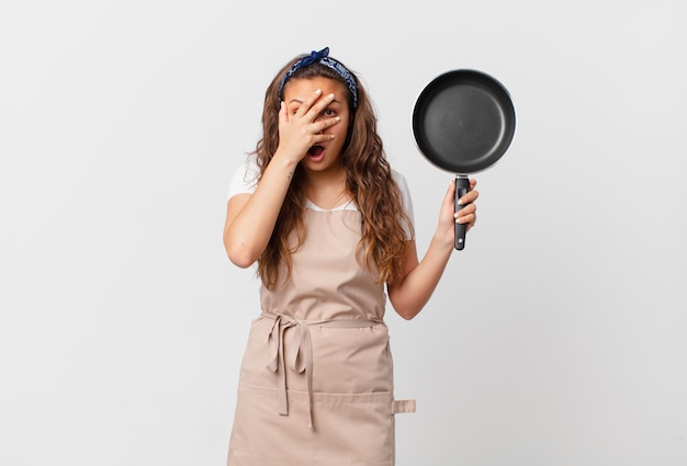 Mujer bonita joven que parece sorprendida, asustada o aterrorizada, cubriendo la cara con el concepto de chef de mano y sosteniendo una sartén