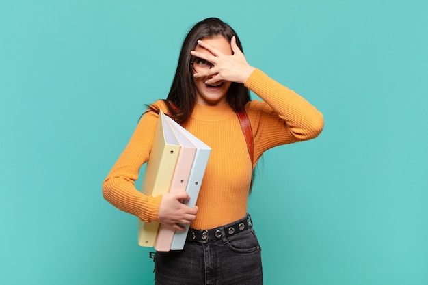 Mujer bonita joven que parece sorprendida, asustada o aterrorizada, cubriendo la cara con la mano y mirando entre los dedos. concepto de estudiante