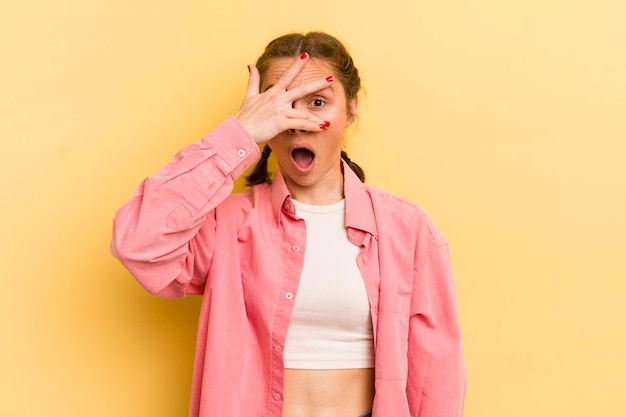 Mujer bonita joven que parece sorprendida asustada o aterrorizada cubriendo la cara con la mano y mirando entre los dedos