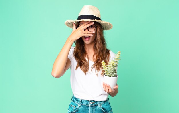 Mujer bonita joven que parece sorprendida, asustada o aterrorizada, cubriendo la cara con la mano con un sombrero de paja y sosteniendo un cactus