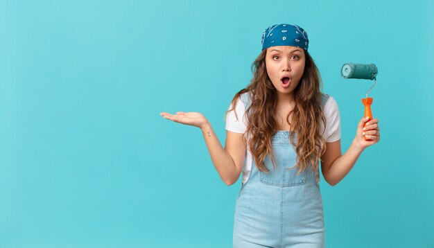 Mujer bonita joven que parece sorprendida y conmocionada, con la mandíbula caída sosteniendo un objeto y pintando una pared