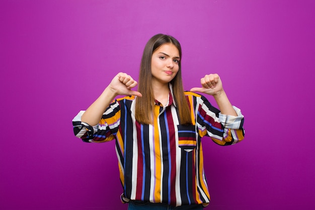 Mujer bonita joven que parece triste, decepcionada o enojada, mostrando los pulgares hacia abajo en desacuerdo, sintiéndose frustrada contra la pared púrpura