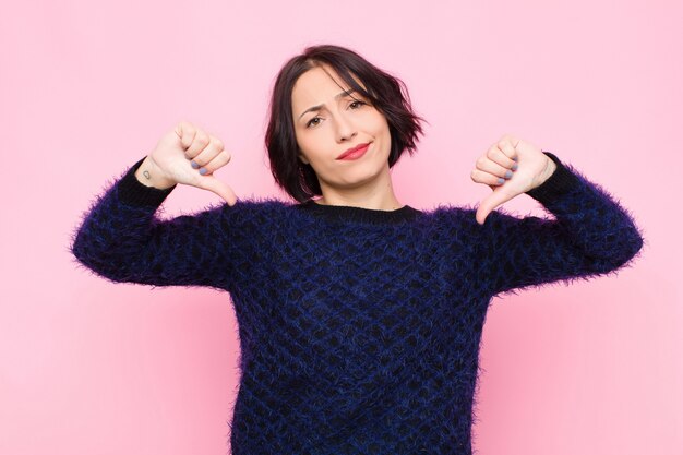 Mujer bonita joven que parece triste, decepcionada o enojada, mostrando los pulgares hacia abajo en desacuerdo, sintiéndose frustrada por la pared rosa