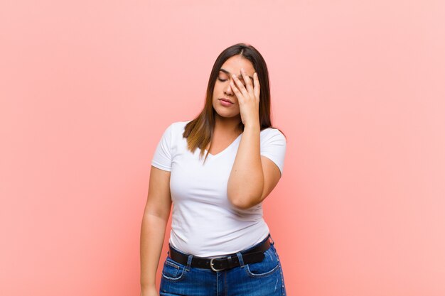 Mujer bonita joven que se siente aburrida, frustrada y somnolienta después de una tarea aburrida, aburrida y tediosa, sosteniendo la cara con la mano contra la pared rosa