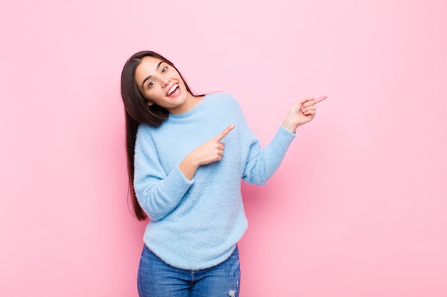 Mujer bonita joven que se siente alegre y sorprendida, sonriendo con una expresión de asombro y apuntando hacia un lado sobre la pared rosa