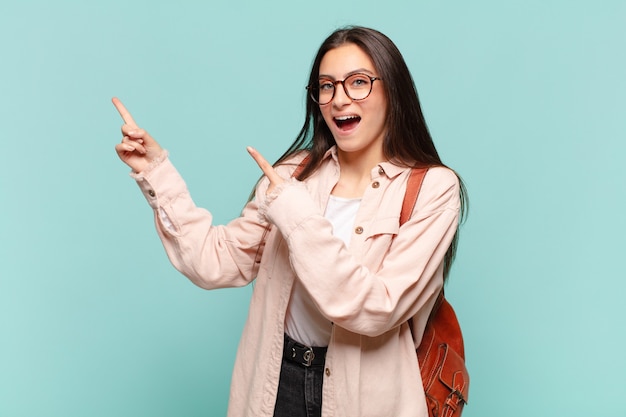 Mujer bonita joven que se siente alegre y sorprendida, sonriendo con una expresión de asombro y apuntando hacia un lado