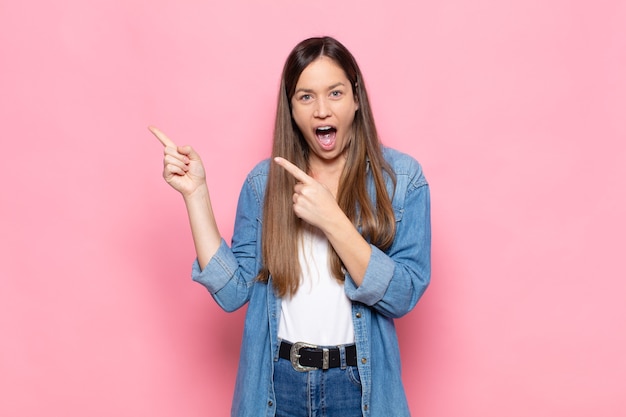 Mujer bonita joven que se siente alegre y sorprendida, sonriendo con una expresión de sorpresa y apuntando hacia un lado