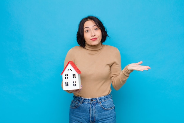 Mujer bonita joven que se siente confundida y confundida, dudando, sopesando o eligiendo diferentes opciones con expresión divertida con un modelo de casa