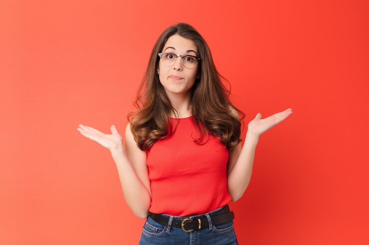 Foto mujer bonita joven que se siente confundida y confundida, dudando, sopesando o eligiendo diferentes opciones con expresión divertida sobre la pared roja