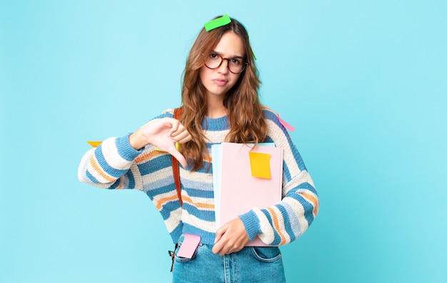 Mujer bonita joven que se siente cruzada, mostrando los pulgares hacia abajo con una bolsa y sosteniendo libros