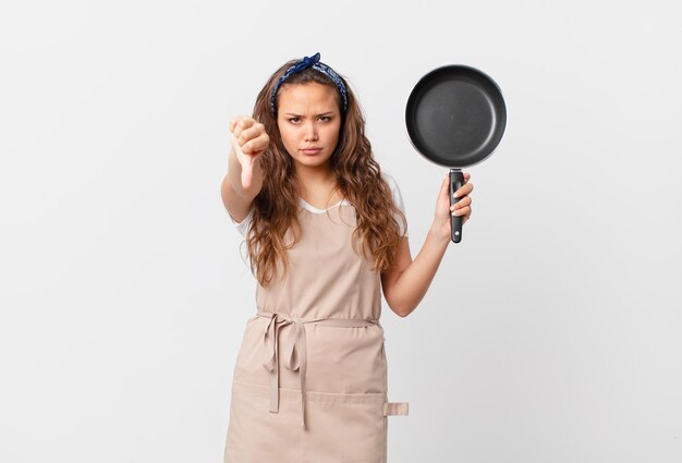 Foto mujer bonita joven que se siente cruzada, mostrando los pulgares hacia abajo el concepto de chef y sosteniendo una sartén