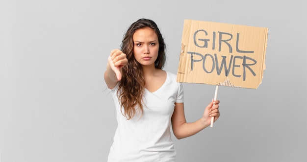 Mujer bonita joven que se siente cruzada, mostrando los pulgares hacia abajo y sosteniendo un cartel de poder femenino