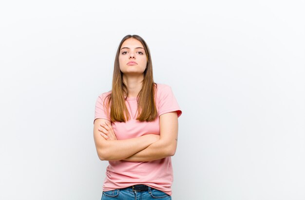 Mujer bonita joven que se siente disgustada y decepcionada, se ve seria, molesta y enojada con los brazos cruzados contra la pared blanca