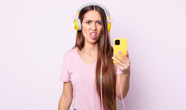 Mujer bonita joven que se siente disgustada e irritada y con la lengua fuera. auriculares y smartphone