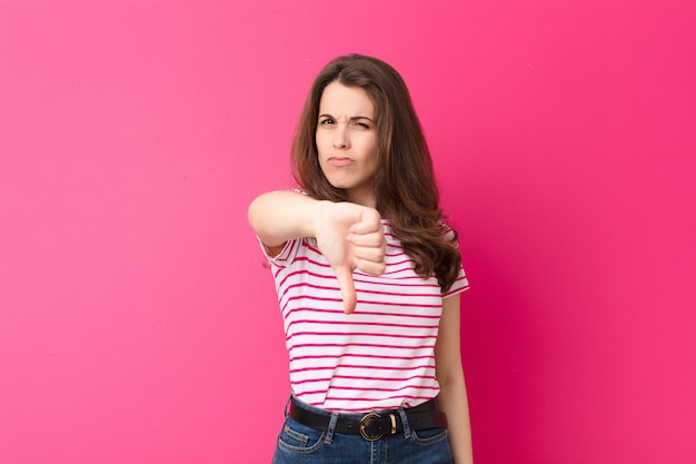 Mujer bonita joven que se siente enfadada, enojada, molesta, decepcionada o disgustada, mostrando los pulgares hacia abajo con una mirada seria contra la pared rosa