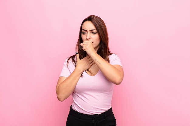 Mujer bonita joven que se siente enferma con dolor de garganta y síntomas de gripe, tos con la boca cubierta contra la pared rosa