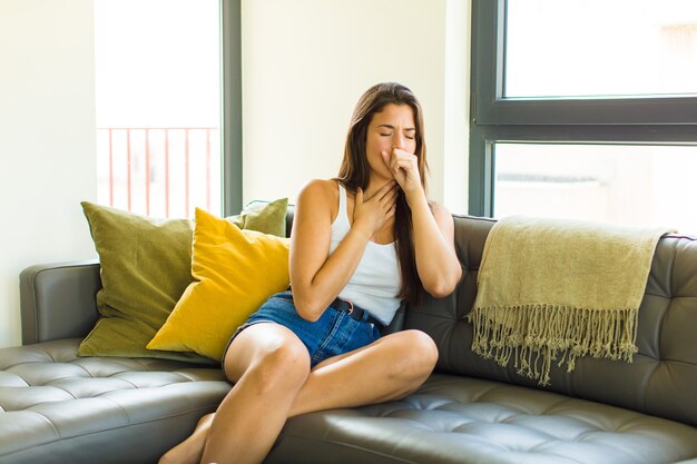 Mujer bonita joven que se siente enferma con dolor de garganta y síntomas de gripe, tos con la boca cubierta