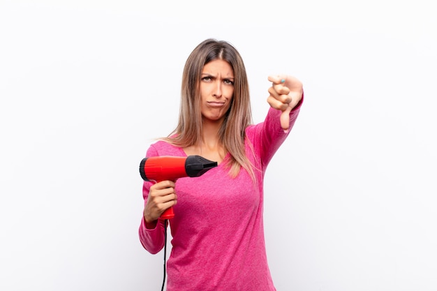 Mujer bonita joven que se siente enojada, enojada, molesta, decepcionada o disgustada, mostrando los pulgares hacia abajo con una mirada seria con un secador de pelo.