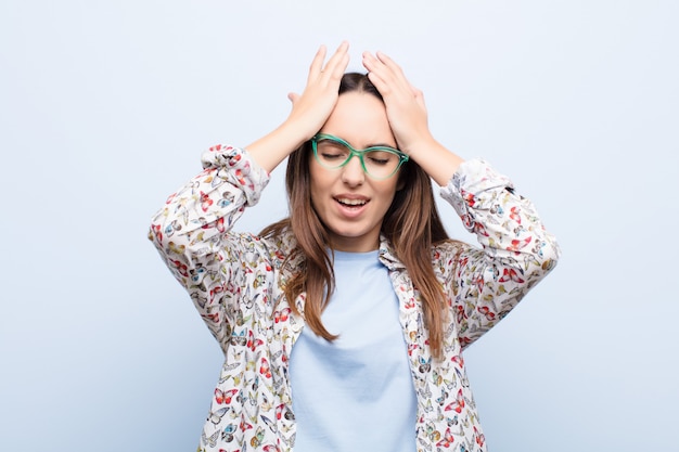 Mujer bonita joven que se siente estresada y ansiosa, deprimida y frustrada con dolor de cabeza, levantando ambas manos contra la pared azul