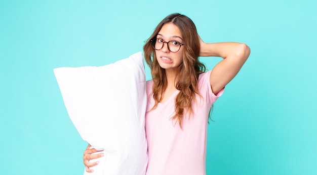 Mujer bonita joven que se siente estresada, ansiosa o asustada, con las manos en la cabeza en pijama y sosteniendo una almohada