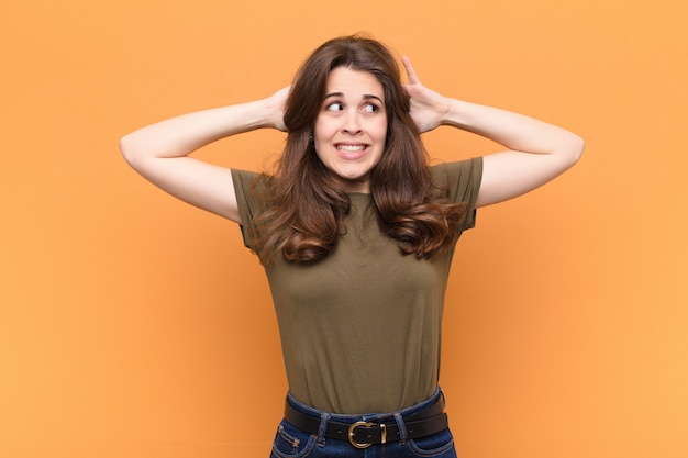 Mujer bonita joven que se siente estresada, preocupada, ansiosa o asustada, con las manos en la cabeza, aterrorizada por error contra la pared naranja