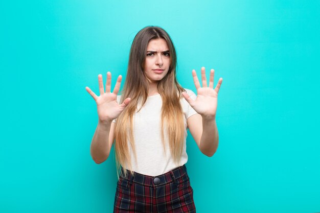 Mujer bonita joven que se siente estupefacta y asustada, temiendo algo aterrador, con las manos abiertas al frente diciendo alejarse sobre la pared azul