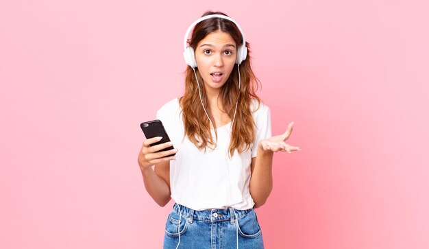 Mujer bonita joven que se siente extremadamente conmocionada y sorprendida con auriculares y un teléfono inteligente