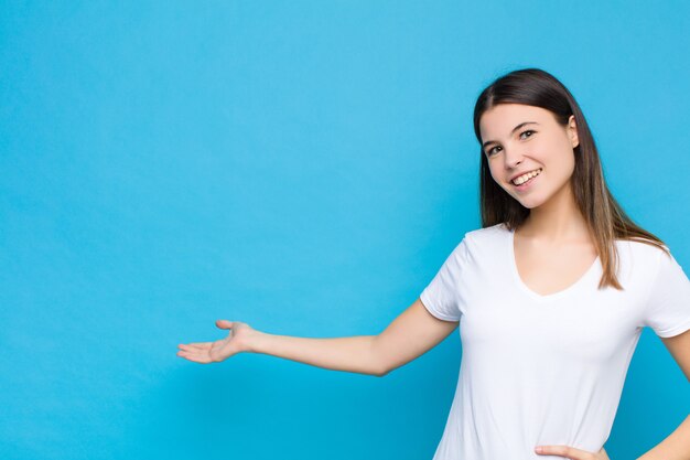 Mujer bonita joven que se siente feliz y alegre, sonriendo y dándote la bienvenida, invitándote a entrar con un gesto amistoso contra la pared azul