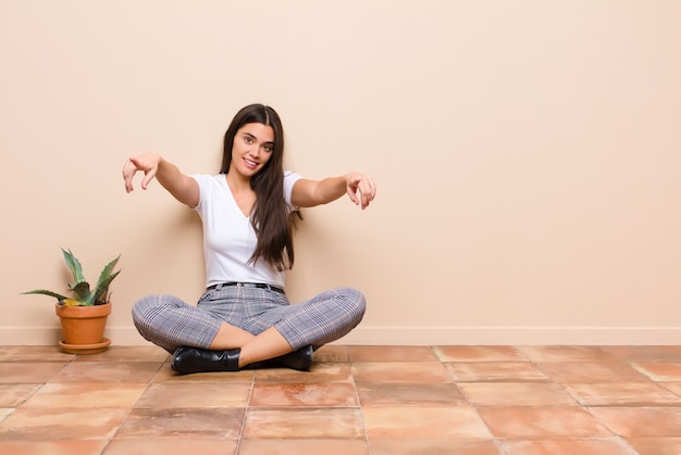 Foto mujer bonita joven que se siente feliz y confiada, apuntando a la cámara con ambas manos y riendo, eligiéndote sentado en el suelo