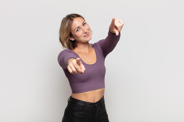 Mujer bonita joven que se siente feliz y confiada, apuntando a la cámara con ambas manos y riendo, eligiéndote