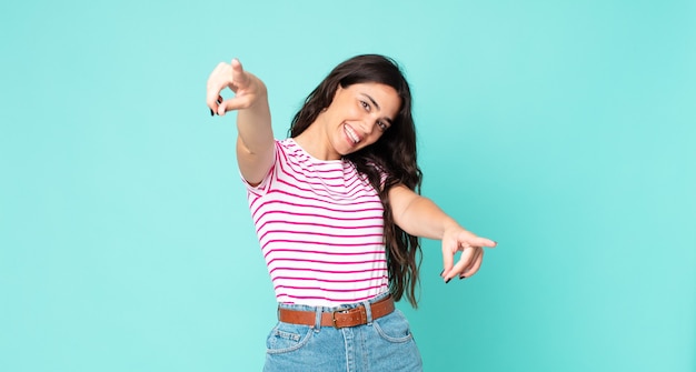 Mujer bonita joven que se siente feliz y confiada, apuntando a la cámara con ambas manos y riendo, eligiéndote