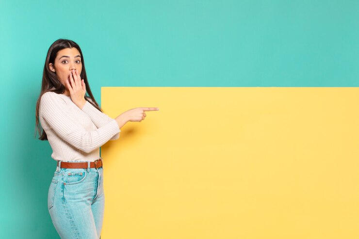Foto mujer bonita joven que se siente feliz, conmocionada y sorprendida, cubriendo la boca con la mano y apuntando al espacio lateral de la copia. copia espacio para colocar tu concepto
