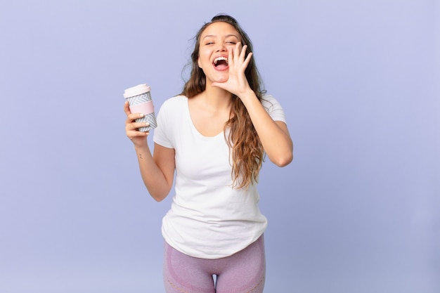 Mujer bonita joven que se siente feliz, dando un gran grito con las manos al lado de la boca y sosteniendo un café