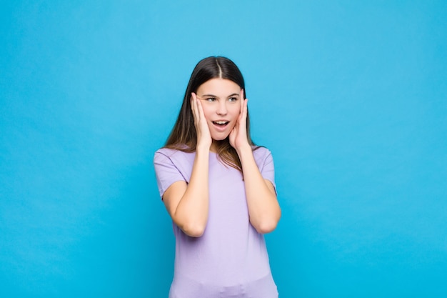 Mujer bonita joven que se siente feliz, emocionada y sorprendida, mirando hacia un lado con ambas manos en la cara contra la pared azul