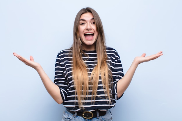 Mujer bonita joven que se siente feliz, emocionada, sorprendida o sorprendida, sonriendo y asombrada por algo increíble contra la suave pared azul