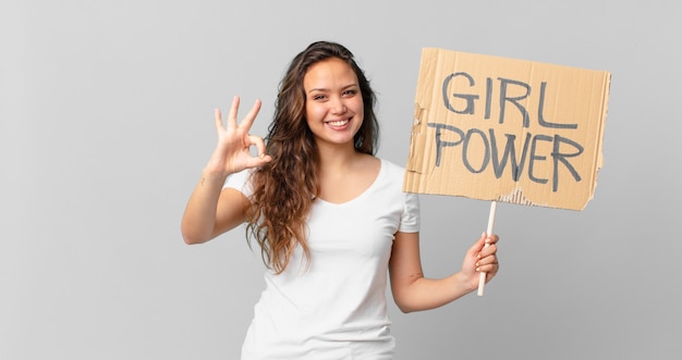 Mujer bonita joven que se siente feliz, mostrando aprobación con un gesto aceptable y sosteniendo una pancarta de poder femenino