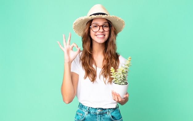 Mujer bonita joven que se siente feliz, mostrando aprobación con gesto bien con un sombrero de paja y sosteniendo un cactus